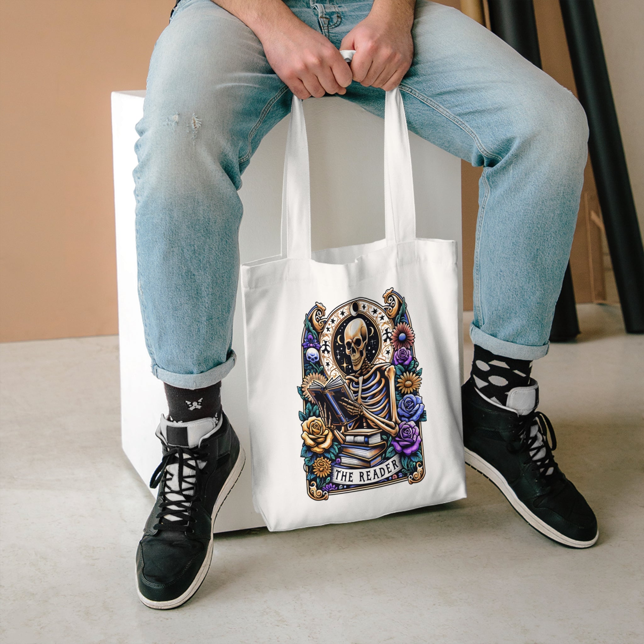 Person holding a tote bag with a skeleton design and 'The Reader' text, wearing light blue jeans and black sneakers.