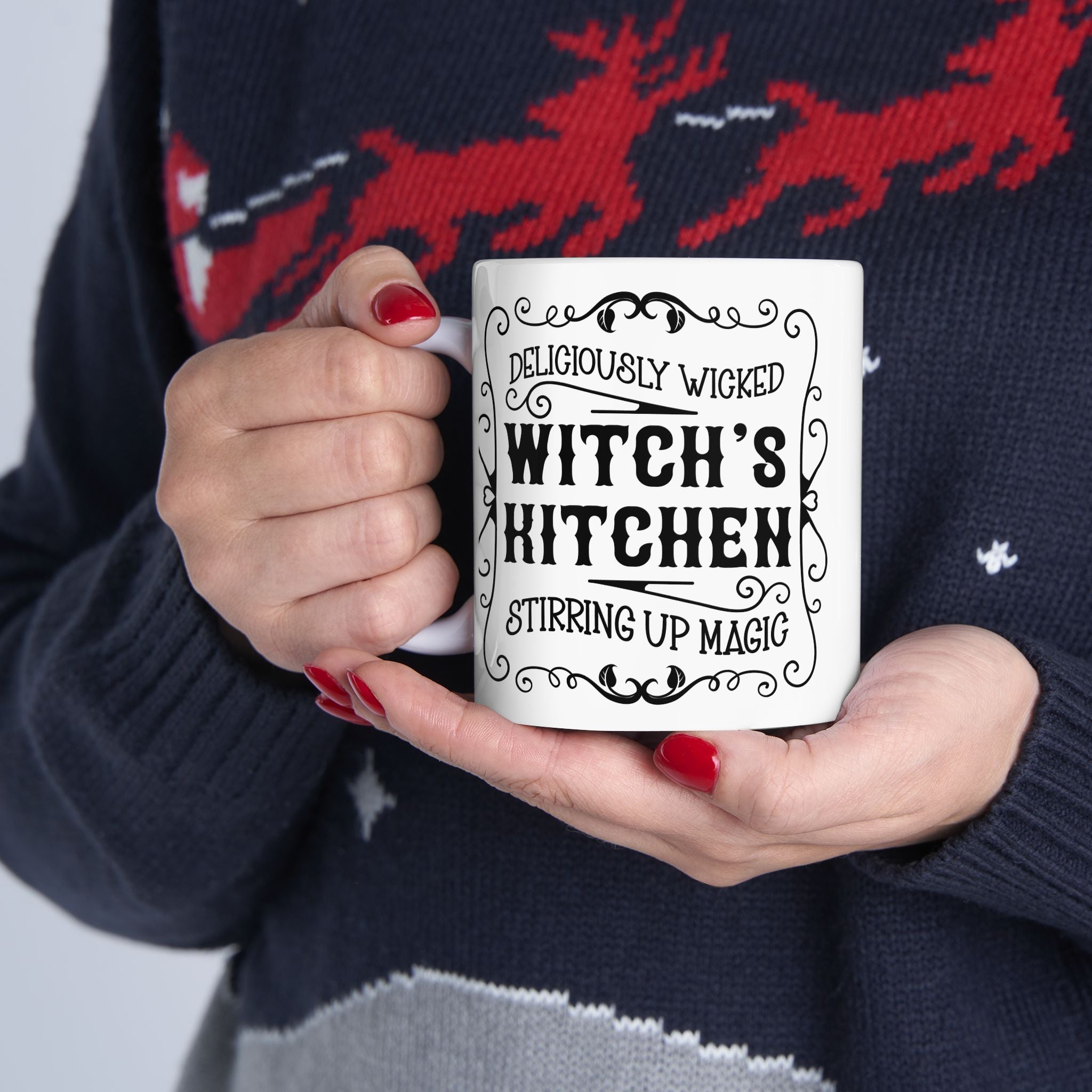 Person holding a mug with 'Deliciously Wicked Witch's Kitchen Stirring Up Magic' text, wearing a festive sweater with reindeer design.